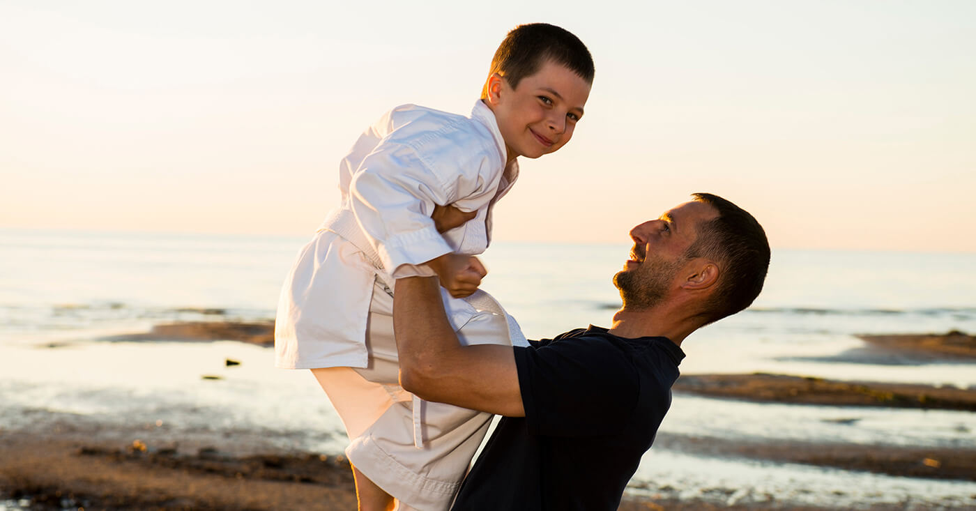 Father holding his son up in the air at the beach. The son is wearing a gi. kids doing karate Father holding his son up in the air at the beach. The son is wearing a gi. kids doing karate