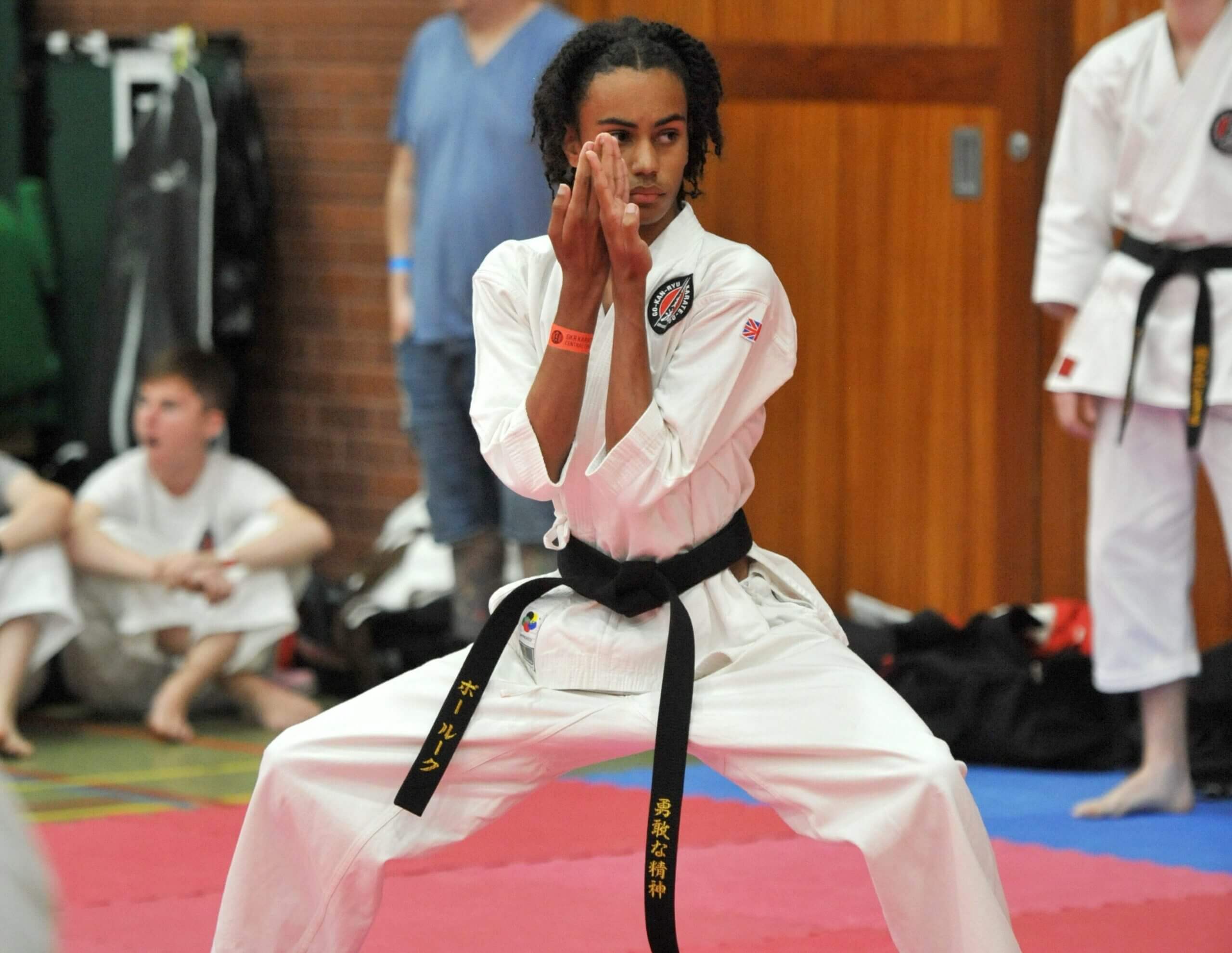 bushido A focused young karateka with long hair is captured in a deep stance, performing a hand technique with intense concentration. Wearing a white gi and a black belt, the practitioner's gaze is directed forward, exuding calm and discipline. In the background, observers and fellow competitors are seated around the edge of the red and blue matted area of the dojo
